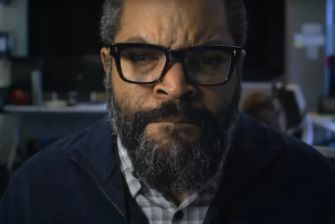 Close-up of a man with a beard and glasses, looking serious against a blurred background.