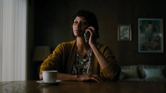 A woman sitting at a table on the phone, wearing a yellow cardigan and looking contemplatively towards the side.