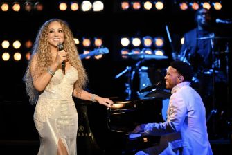 Mariah Carey performing on stage, singing while accompanied by a pianist, with bright stage lights in the background.