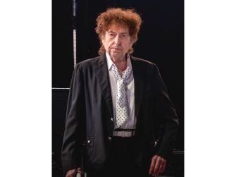 Bob Dylan standing on stage with a serious expression, wearing a black suit and a white shirt with a polka-dot tie.