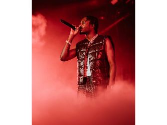 Rapper Lil Tjay performing on stage with a microphone, surrounded by red lighting and smoke.