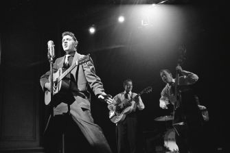 Elvis Presley performing on stage with a guitar, accompanied by other musicians in a vintage black-and-white setting.