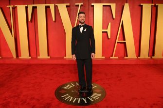 A formally dressed man standing on a red carpet with the words "UNITY FALL" in the background at an event.