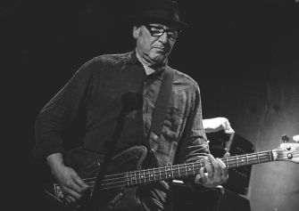 A black and white image of a musician playing a bass guitar on stage, wearing glasses and a hat.