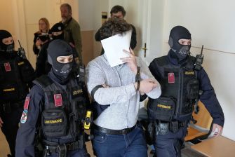 A man wearing a paper mask to hide his identity is escorted by armed police officers in a courtroom setting.