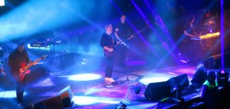 A live performance by New Order featuring band members on stage with lighting effects and instruments.