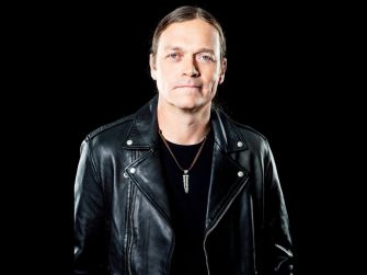 A man in a leather jacket stands against a black background, looking directly at the camera with a serious expression.