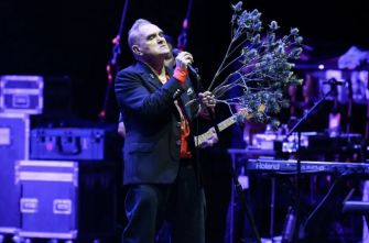 Morrissey performing on stage while holding flowers, surrounded by musical equipment.