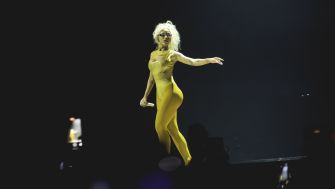 A performer in a yellow outfit striking a dynamic pose on stage, surrounded by audience members with their phones raised.