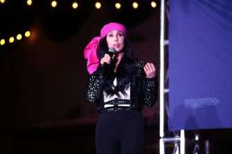 Cher speaking on stage, wearing a black outfit and a pink headscarf, holding a microphone.