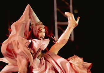 A performer in a dramatic costume, featuring a crown and flowing garments, passionately engaging with the audience during a performance.