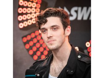 A portrait of singer Lauv during an interview, looking contemplative amidst a backdrop of stage lights.