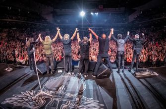 A group of musicians on stage with their arms raised, facing a large audience during a concert.