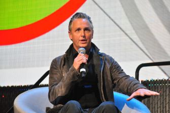 Mike McCready of Pearl Jam playing guitar while seated, wearing a brown leather jacket and focused on his instrument.