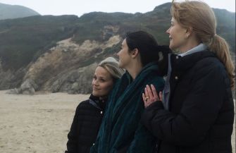 Three women standing on a beach, looking towards the horizon, wearing winter attire against a scenic coastal backdrop.
