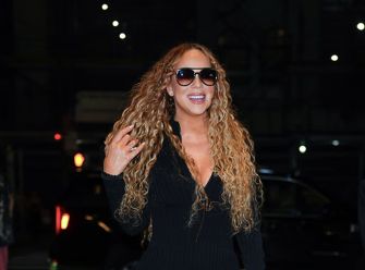 Mariah Carey smiling and posing outdoors while wearing sunglasses and a black outfit.
