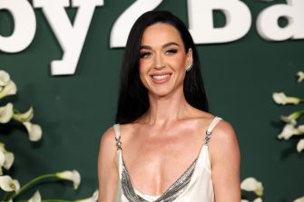 Katy Perry smiling at an event, wearing a stylish outfit against a floral background.