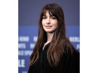 Anne Hathaway at a promotional event for her upcoming film, wearing a black outfit and posing against a blue backdrop.