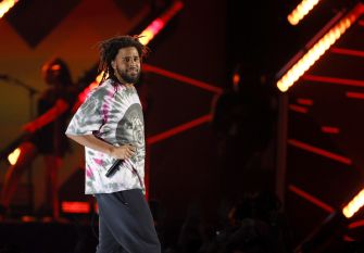 J. Cole performing on stage, wearing a tie-dye shirt and looking toward the audience.