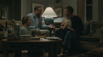 A dinner gathering scene featuring four actors in a cozy living room setting, with a lamp illuminating the space.
