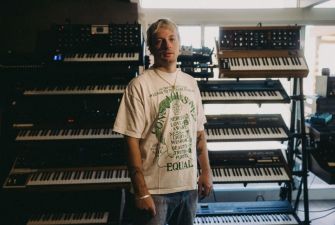 Kid Harpoon standing in a studio surrounded by various synthesizers and musical equipment.