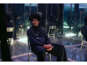 Labrinth sitting on a chair in a reflective space, looking contemplative while holding a snack.