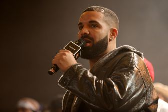 Drake speaking into a microphone during an event, wearing a leather jacket.