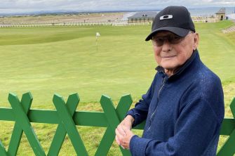 James Tolkan standing outdoors near a golf course, wearing a dark cap and a blue sweater.