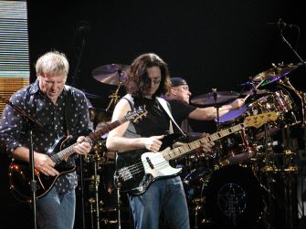 Rush performing on stage with Alex Lifeson on guitar, Geddy Lee on bass, and Neil Peart on drums.