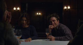 A scene from the film "The Drama," featuring two characters seated at a table with drinks, engaged in conversation.