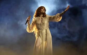Florence Welch performing on stage in a flowing white outfit, holding a microphone under dramatic lighting.