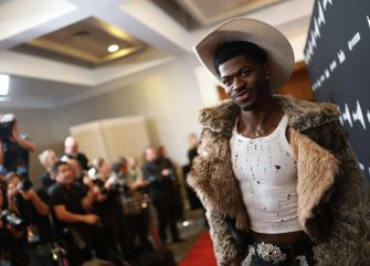 Lil Nas X posing on the red carpet wearing a cowboy hat and a fur coat, surrounded by photographers.