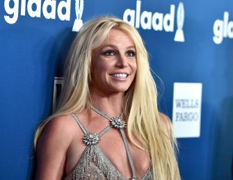 Britney Spears smiling at an event, with a blue background featuring sponsor logos.