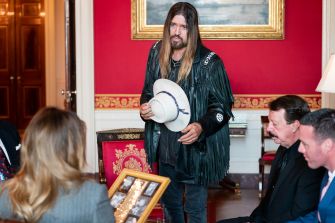 Billy Ray Cyrus stands in a formal setting, holding a hat and speaking to a group of people seated nearby.