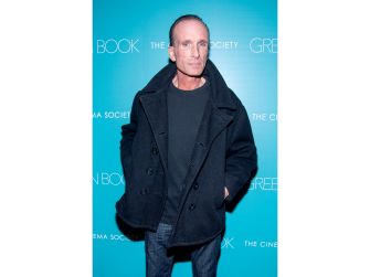 Peter Greene at a film event, wearing a black coat and standing in front of a teal backdrop with text.