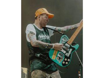 Chad Gilbert performing on stage with a guitar, wearing a cap and a t-shirt, surrounded by musical equipment.