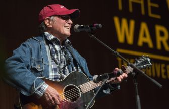 Country Joe McDonald performing on stage with an acoustic guitar, wearing a cap and denim jacket.