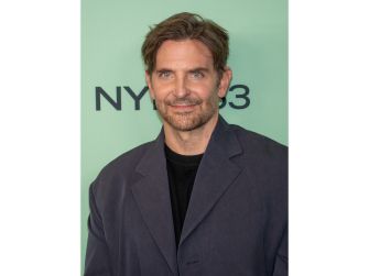 Bradley Cooper at an event, smiling and wearing a dark jacket against a light green background.