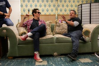 Two men sitting on a couch, engaging in conversation while wearing casual attire, with a patterned wall in the background.