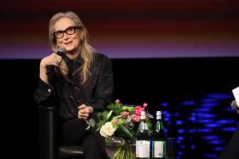 Meryl Streep speaking at an event, sitting with a floral arrangement and bottles of water.