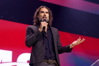 Russell Brand speaking on stage at an event, holding a microphone.