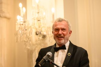 Actor Sam Neill smiling at an event, wearing a tuxedo, with a chandelier in the background.
