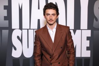 A man in a brown suit poses for a photo against a backdrop featuring bold text.