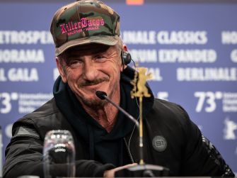 Sean Penn at a press conference, wearing a camouflage hat and jacket, smiling while speaking into a microphone.