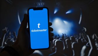 A person holding a smartphone displaying the Ticketmaster logo, with a blurred crowd at a concert in the background.