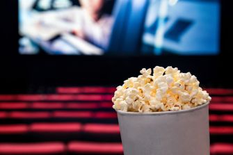 A close-up of a cup of popcorn in a nearly empty movie theater with a blurred screen in the background.