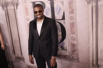 Kanye West, wearing a black suit and sunglasses, poses for a photo at an event with a decorative backdrop.