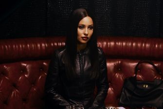 A woman sitting on a red leather couch, wearing a black leather jacket, with a serious expression. A handbag and drinks are placed on the table in front of her.