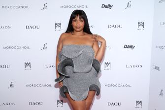 A woman in a sleek, gray strapless dress poses confidently at a red carpet event with promotional logos in the background.