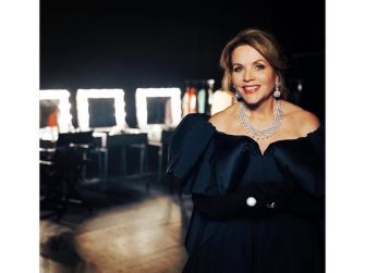 Renée Fleming smiling in a backstage setting, wearing an elegant black dress and sparkling jewelry.